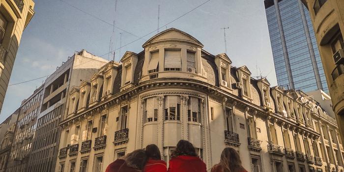 4 mujeres, en un balcón,  vestidas de rojo y de espaldas, frente a edificios antiguos